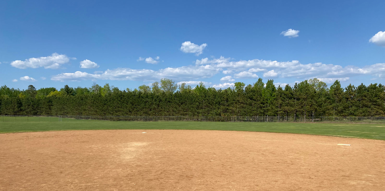 Empty baseball field with blue sky, white clouds, and lush trees in background.