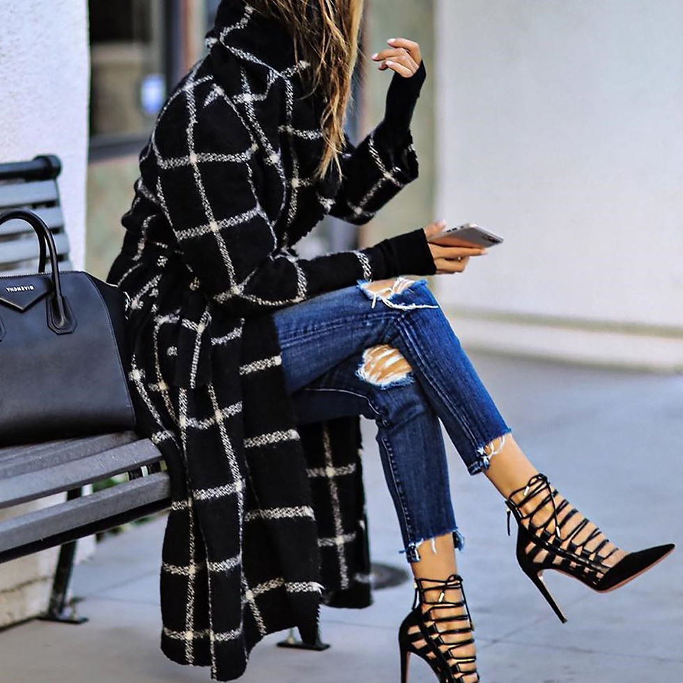 Woman sitting on a bench wearing a long black and white checkered belted overcoat, ripped blue jeans, and black strappy heels, holding a phone.