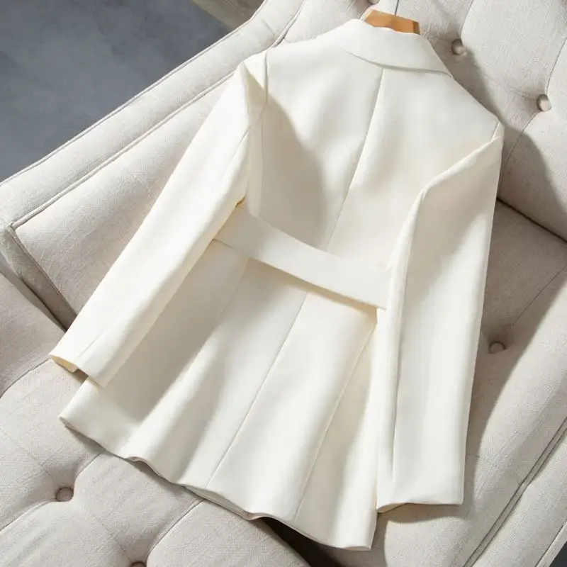 縮圖：White women's blazer displayed on a beige tufted chair, showing the back design with a waist belt detail and structured tailoring.