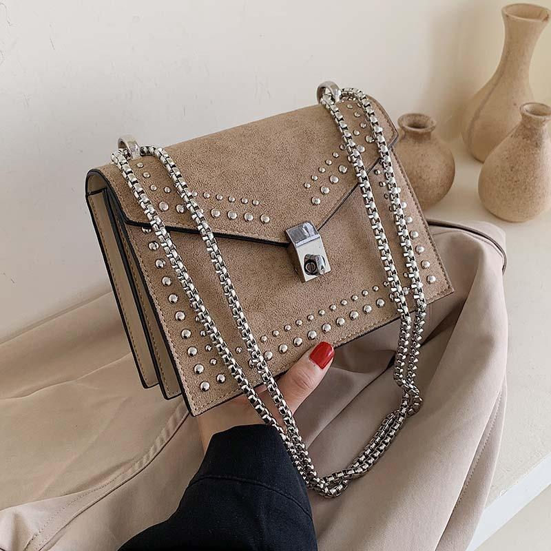 Tan scrub leather shoulder bag with silver chain straps, metal rivet detailing, and a lock clasp, displayed against a neutral background with ceramic vases.
