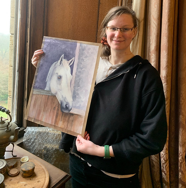 Artist Pippa Hobbs smiling holding a finished commission piece of a white horse in a stable, painted using watercolours
