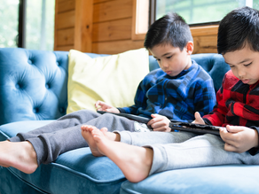 Two children sitting together on a couch but completely absorbed in their individual handheld devices, demonstrating how screen time can reduce face-to-face social interaction and connection between siblings or friends.
