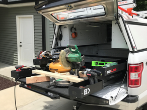 Pickup truck tailgate workbench loaded with saws, drill, lumber and tools outside a garage, with BedSlide text visible