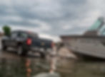Pickup truck launching a white boat in the water, under a cloudy sky. The scene is calm and overcast. BW Trailer Hitch