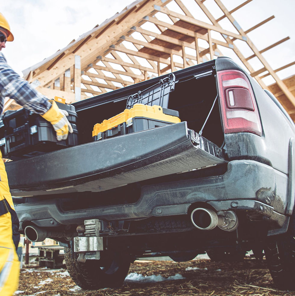 A person in plaid and yellow overalls loads toolboxes into a truck bed at a construction site. Wooden frame structure in the background.
