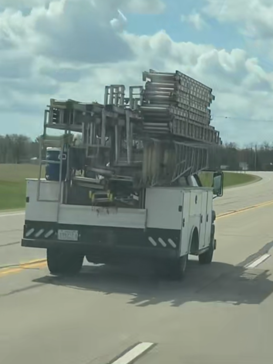 Utility truck hauling stacked metal ladders on a highway under cloudy skies.