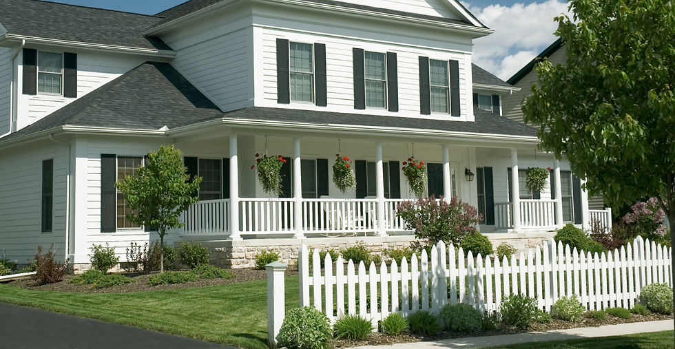 White house with black shutters, front porch, hanging plants, white picket fence, green lawn, trees, and sunny blue sky. Peaceful suburb scene.