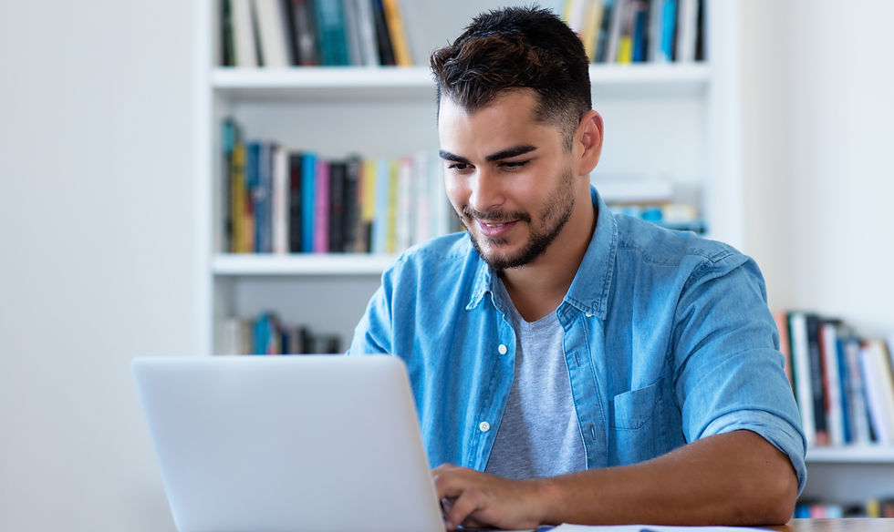 Man in denim shirt smiling while using a laptop in a room with a bookshelf. Bright, relaxed atmosphere with a focus on his expression.