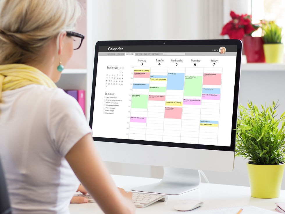 Woman in white shirt views colorful calendar on desktop computer in bright office. Green plant and pink folders nearby on desk.