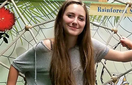 Smiling girl in a gray shirt stands in front of a rope net with greenery. Background has a sign reading "Rainforest" and a red flower.