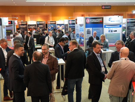 Attendees networking and visiting exhibitor booths at the PIC International Conference 2024 in Brussels. The image shows a busy conference hall with professionals engaged in conversation, surrounded by displays from various companies and organizations in the photonics and semiconductor industries.