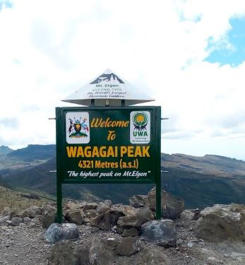 Sign reading "Welcome to Wagagai Peak, 4321 Metres (a.s.l), The highest peak on Mt. Elgon" set against a mountain landscape and cloudy sky.