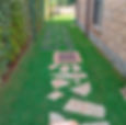 Stone path with irregular shapes and square slabs on green arabic grass, flanked by a brick wall and hedge. Bright, vibrant backyard scene.