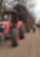 Red Kubota tractor pulling a large load of twigs on a dirt road. A person walks alongside, with trees and a second tractor in the background.