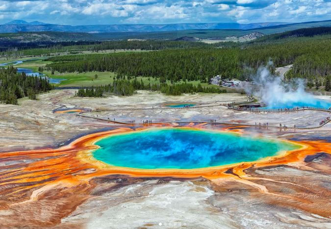 Vibrant geothermal pool with blue center, surrounded by orange and yellow rings. Boardwalk with people nearby; forested backdrop under blue sky.