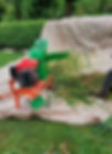 Green and red agricultural machine chopping grass on a tarp-covered lawn. Bushes and trees in the background. Worker partially visible.