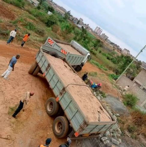 A large truck stuck in a dirt road, surrounded by people. Overcast sky, rural setting with greenery and buildings in the background.