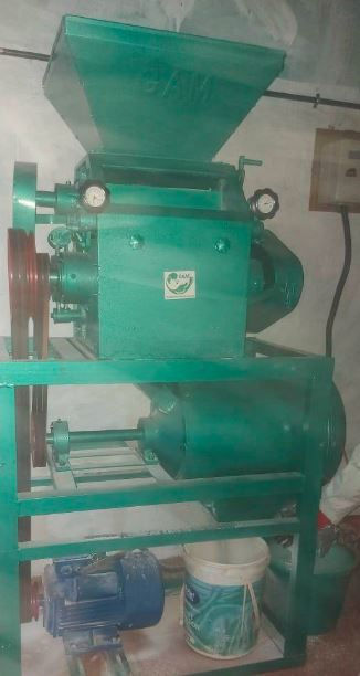 Green industrial machine with gauges and a hopper, set in a workshop. A bucket is underneath. The scene is dimly lit.