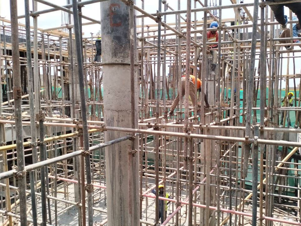 Construction site with workers in hard hats and safety gear navigating metal scaffolding. Background shows a partly cloudy sky.