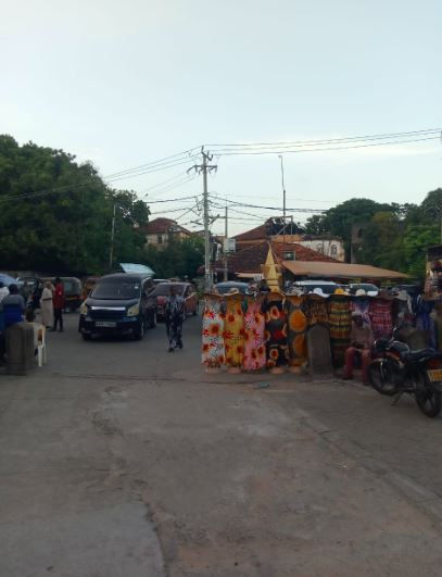 Mombasa Old town Street market with colorful fabric stalls and people walking. Parked cars and trees in the background. Calm evening atmosphere.