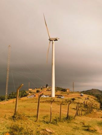 Wind turbine on a grassy hill with dark clouds. Several cars and buildings in the background. Fence leads to the horizon, creating a serene mood.