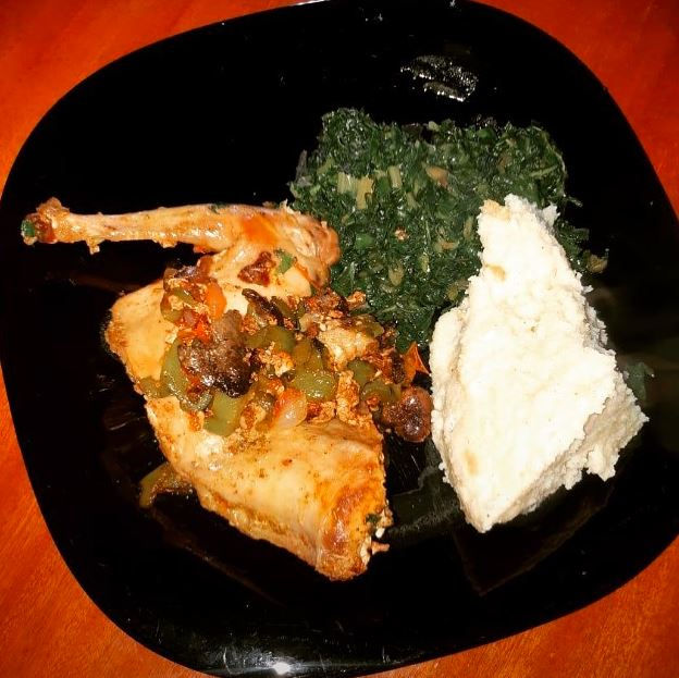 A black plate with stewed chicken, greens, and a scoop of white ugali on a wooden table, showcasing a warm, homemade meal.