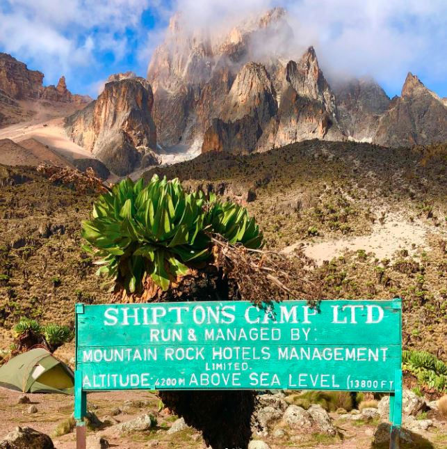 Green sign reads "Shiptons Camp Ltd" against mount Kenya backdrop with plants, blue sky, and clouds. Rugged, adventurous setting.