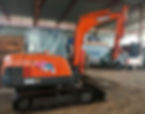 Orange Doosan excavator in a warehouse, parked beside a white truck. Industrial setting with metal roof and stacked materials in the background.