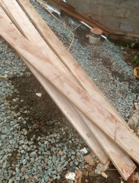 Wooden planks lie on gravel, alongside a paint can and scattered wires, in an outdoor setting. The mood is rustic and industrial.