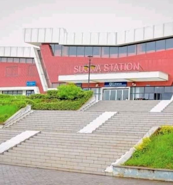 Red building with "SUWON STATION" signage, wide steps leading to entrance, greenery on both sides, overcast sky.