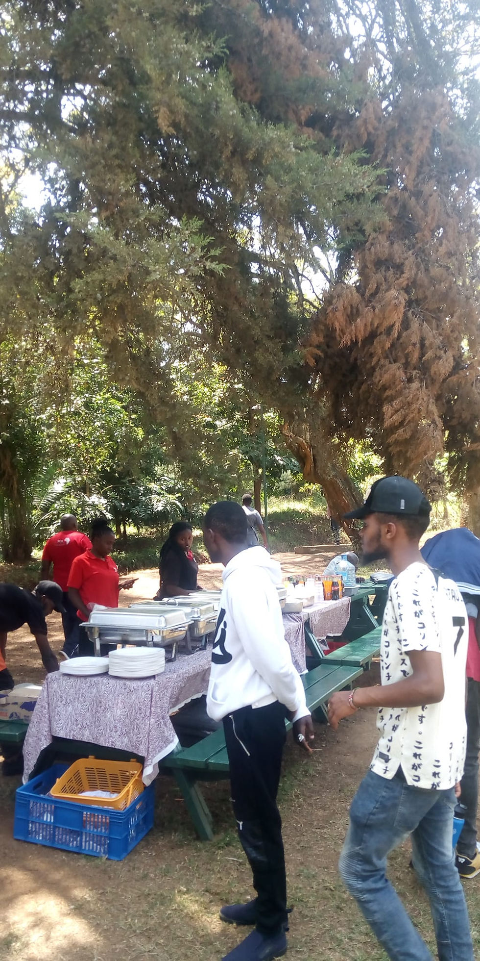 People gather around a picnic table at karura forest with food trays outdoors under large trees. Casual setting with vibrant shirts and relaxed mood.