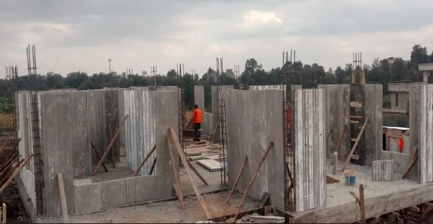 Precast Concrete walls under construction with wooden supports. A person in an orange shirt works on site. Cloudy sky, trees in background.