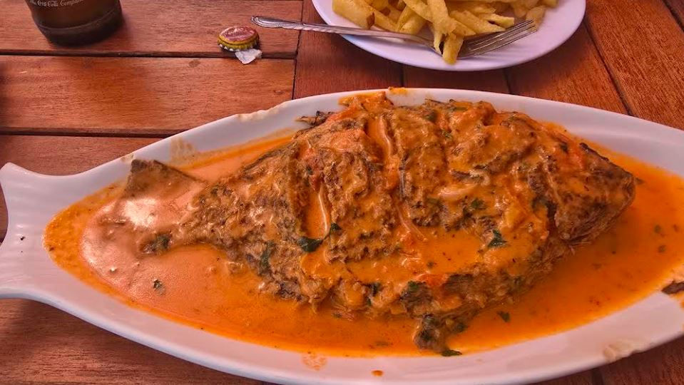 Whole fish in orange sauce on a white platter, with fries on a plate behind it, served on a wooden table.