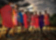 A group of people in colorful traditional attire stand outdoors under a dramatic cloudy sky at dusk, holding staffs and appearing cheerful.