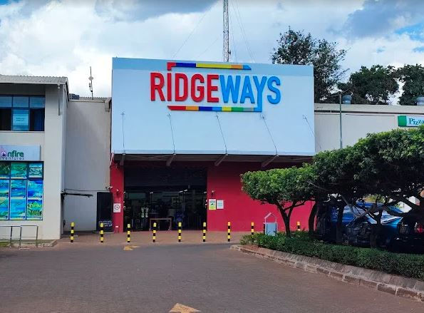 Shopping center entrance with "RIDGEWAYS" sign in bold colors, surrounded by trees and yellow bollards. Clear skies and a relaxed atmosphere.
