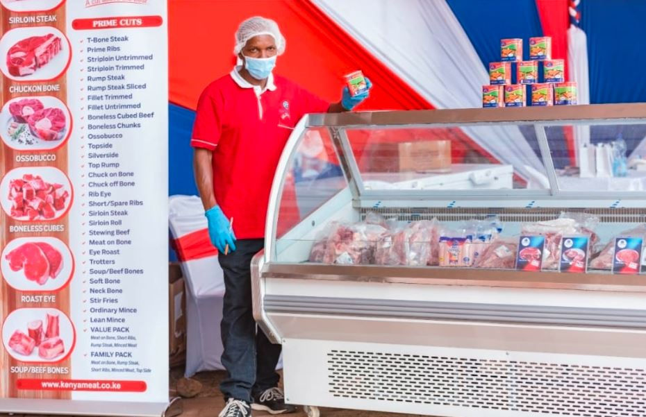 Man in red shirt and mask stands by a meat display in a market. Meat cuts listed on sign. Red, white, and blue drapes in background.