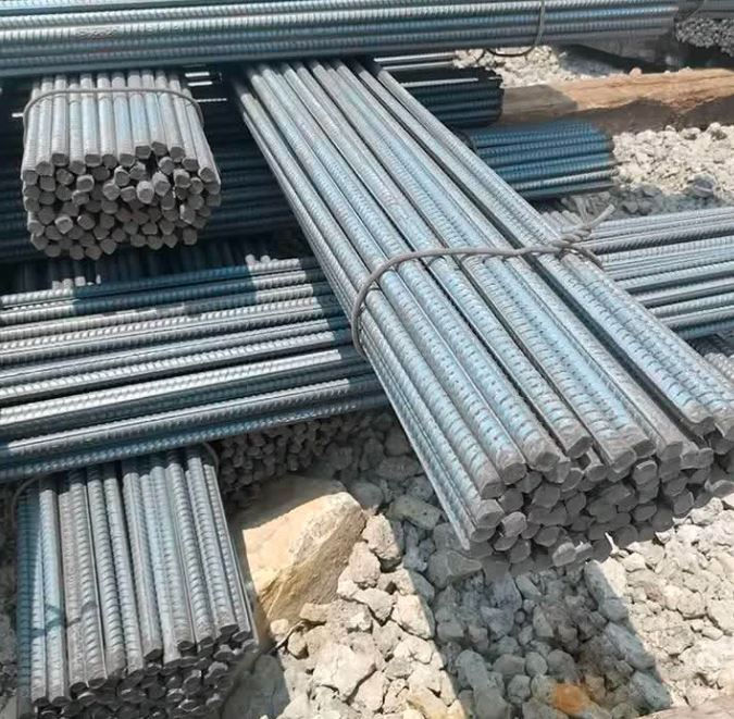 Stacks of steel reinforcing bars are arranged on a rocky surface under bright sunlight. The bars are bundled and parallel.