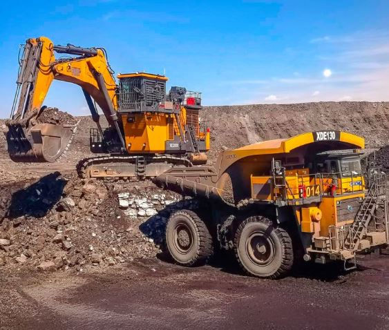 Yellow excavator loads soil into a large dump truck in a quarry. Clear blue sky and rocky terrain in the background.
