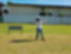 Person in white shirt and jeans swings a golf club on a grassy field. Blue sky and metal building in background. Sign with text visible nearby.