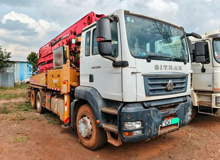 White Sitrak truck with red crane parked on dirt ground. Blue skies with clouds above. License plate partially visible, building in background.