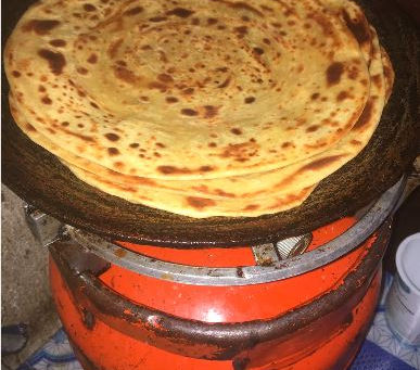 Stack of golden flatbreads cooking on a round griddle atop an orange stove in a kitchen, with a warm homemade feel.