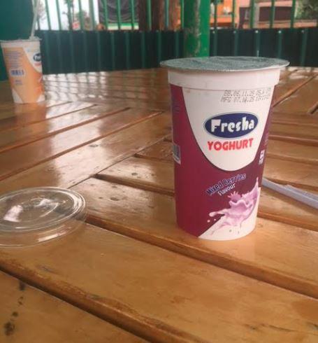 A cup of Fresha yoghurt on a wooden table, with another cup in the background. The setting appears outdoor, with green railings visible.