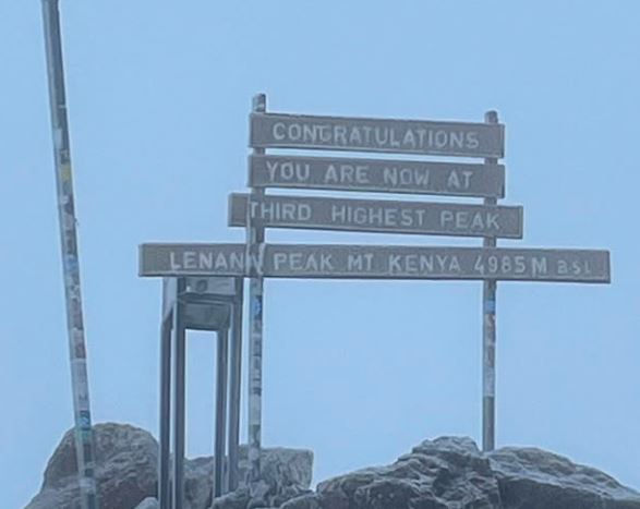 Sign with "Congratulations, You are now at Third Highest Peak, Lenana Peak, Mt Kenya 4985m asl" on rocky, foggy mountain setting.