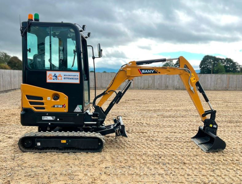 Yellow mini excavator on dirt ground, with trees and wooden fence in the background. Logo on side reads Willow Plant 07714932321. Cloudy sky.