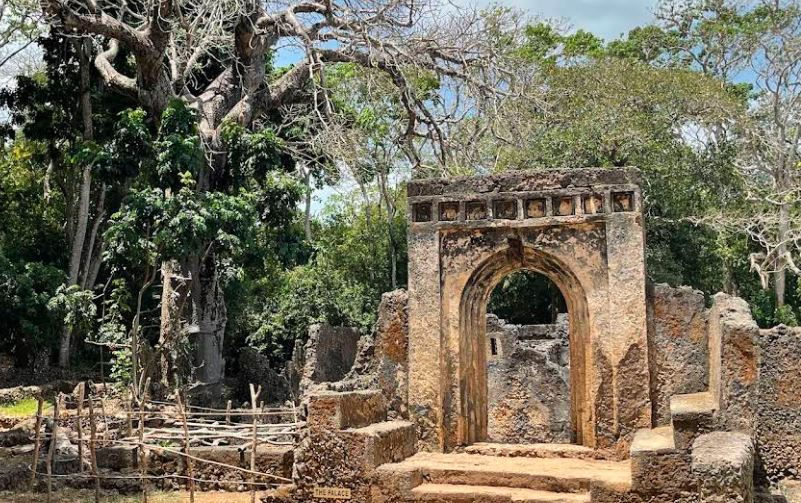 Gede Ruins Ancient stone ruins with arched entrance, surrounded by lush green trees and a clear sky. A wooden fence appears on the left. Mood is tranquil.