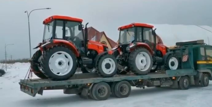 Two red tractors on a flatbed truck in a snowy area. A house and streetlights are visible in the background. The mood is calm.