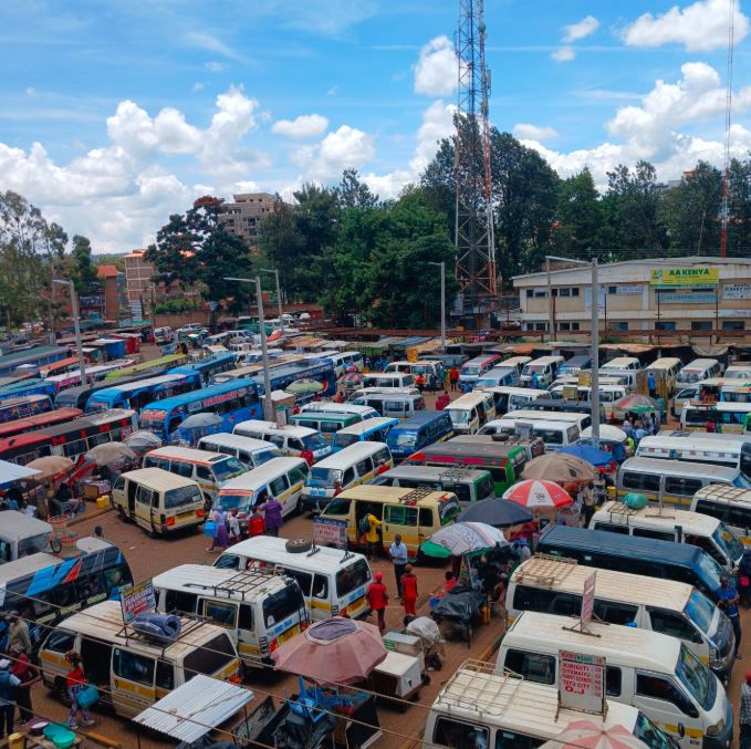 A busy bus terminal filled with colorful vans and people under umbrellas on a sunny day. A few trees and buildings are visible in the background.
