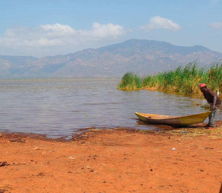Man with a red cap pulls a yellow boat on a lake jipe shore. Background shows mountains, reeds, and blue skies. Calm and serene setting.