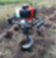 A red and black soil auger drilling a hole in a grassy field, surrounded by loose soil. The mood is industrious and outdoorsy.