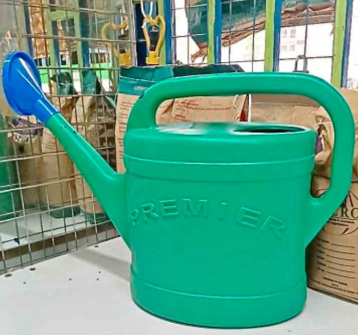 Turquoise watering can with "PREMIER" embossed on side, sits on white surface. Background has bags and grid structure. Bright setting.